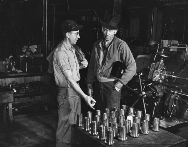 Illustration art of two men in work clothes standing on a factory floor by a turret lathe and work benches talking about newly manufactured machine parts laid out on a table. One man, the machine operator with billed cap and glasses, is pointing at the parts to show the other, who is in a fedora and looking down as he listens. The latter has a Victory-style lunch box under his arm.