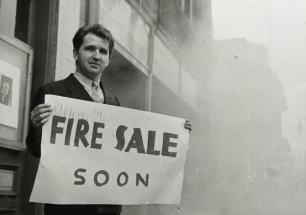 Sid dressed in a suit and tie standing outdoors holding a sheet of paper that reads: "Fire Sale Soon" in front of an unknown building which during a fire. There is smoke in the background. A portrait photograph is hung on the facade of the building just behind Sid. 