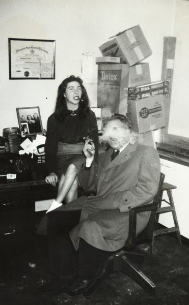 Sid and a woman sitting and smoking in an office. Sid is sitting in a chair wearing an overcoat. His face is obscured by the smoke he is exhaling. The woman is sitting on the desk behind Sid, and she has a cigarette holder in her mouth. On the wall is hanging a certificate from the Wisconsin State Board of Pharmacy. 