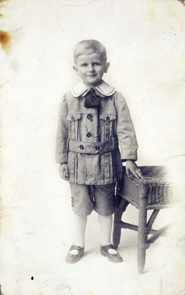 Full-length, vignetted portrait of young John Boyum, Sid's brother,  standing next to wicker furniture. 
