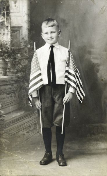 Full-length studio portrait in front of a painted backdrop of a young Sid Boyum holding two U.S. Flags.