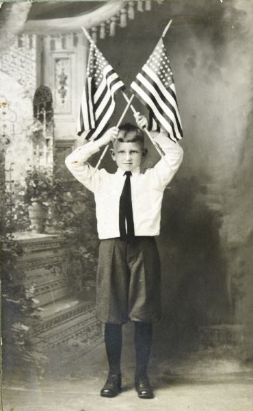 Full-length portrait in front of a painted backdrop of a young Sid holding two U.S. flags crossed above his head. 
