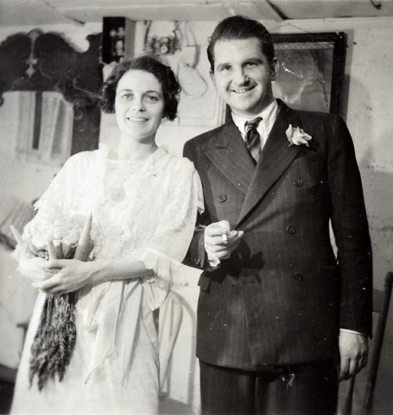 Three-quarter length wedding portrait of Sid and Margaret. They are wearing wedding attire, and Margaret is holding a bunch of carrots and a cauliflower head. Sid is holding a cigarette.