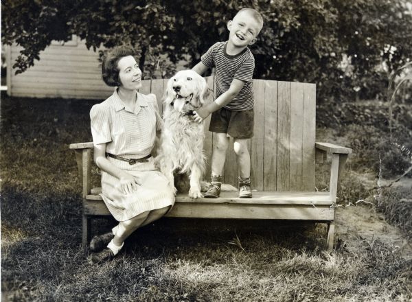 Outdoor portrait of Sid's wife, Margaret and young Steve Boyum sitting with the family dog, Duke, on a wooden bench.