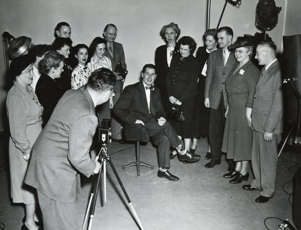 Sid standing behind a tripod taking photographs. A  man is sitting in the center, surrounded by a group of men and women. 