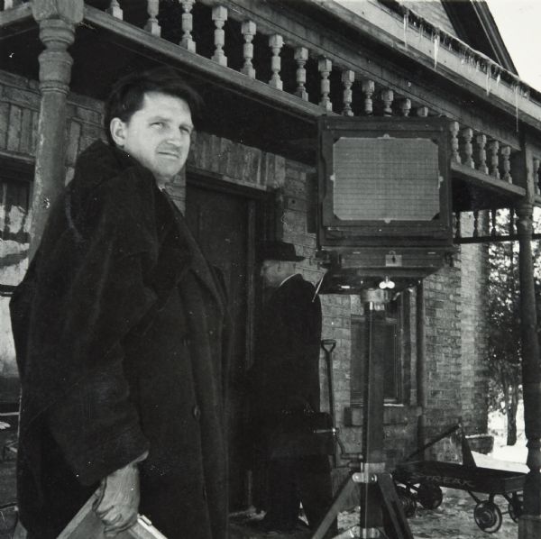 Portrait of Sid with an 8x10 view camera in front of a building in the winter. Sid is wearing a coat and a dark cloth over his shoulder and holding an 8x10 film holder. A man holding a black bag is standing behind Sid near the doorway. On the porch is a shovel and a Streak coaster wagon.