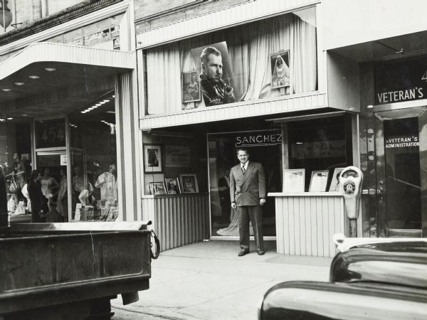 View from street of Sid standing on the sidewalk in front of the Sanchez Photo portrait studio at 11 North Pinckney Street. A large photograph of Sid smoking a pipe is displayed in a window above him on the second floor.