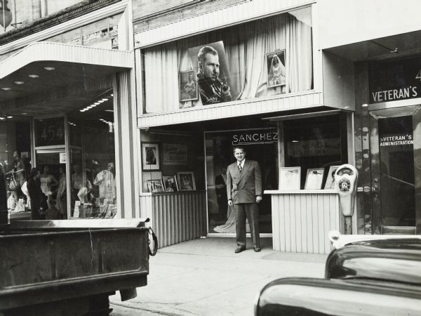 View from street of Sid standing on the sidewalk in front of the Sanchez Photo portrait studio at 11 North Pinckney Street. A large photograph of Sid smoking a pipe is displayed in a window above him on the second floor. 