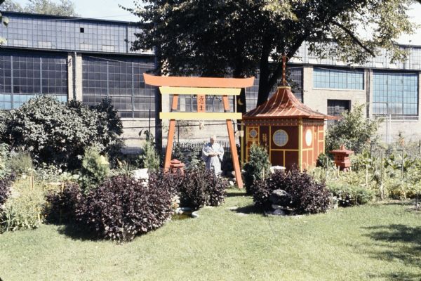 View across lawn towards Sid, standing and wearing a Japanese kimono and near the large arch, the Japanese Tea Pavilion and other lantern sculptures in his backyard. Madison-Kipp Corporation is in the background.