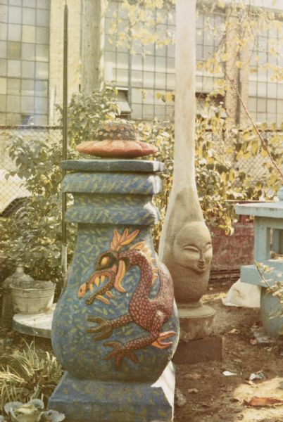 Large concrete-cast sculpture identified as "Blue Dragon Urn" in Sid's backyard. It is painted blue with an Asian styled dragon in red wrapped around it. A blue and red flower acts as a lid, above the neck, tiered like a pagoda. The urn measures 66" x 30" x 28". There are other sculptures surrounding the urn, and Madison-Kipp Corporation is in the background.