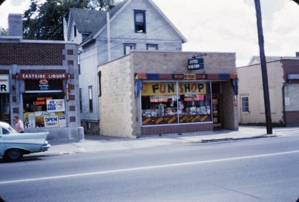 View across street towards Otto's Newstand and Fun Shop located at 2045 Atwood Avenue. There are neon signs for "Otto's News Stand" and "Tobacco Novelties" in the storefront window. Across the alley on the left is Eastside Liquor with a sign that reads "Open Today" and "Ice Cold Beer."