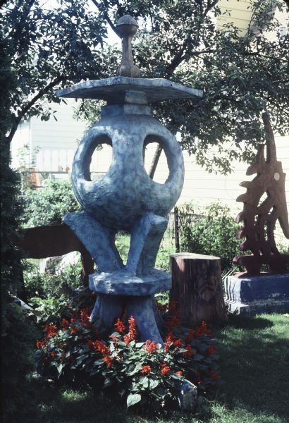 Tall cast-concrete sculpture identified as "Blue Tripod" in Sid's backyard near a fence. It measures 104" x 31" x 54". An abstract fish-like form identified as "African-Influenced Sculpture" is in the background; it measures 94" x 34" 11". 