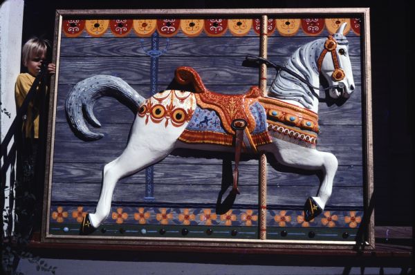 Large, plastic molded plaque of a carousel horse mounted on a decorated, wooden background. The relief, supported by a child on the stairs of Sid's house, is of a white horse with gray man, equipped with a red saddle trimmed in orange, blue saddle blanket and ornate caparison. 