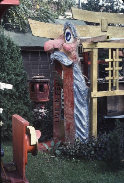 Unpainted wooden Japanese torii gate and a side view of sculptural arch of a standing old man in Sid's backyard. The man, with a prodigious nose, big blue eyes, ruddy complexion and dressed in a gray toga, rests his face and hands on an brown painted arch inscribed with the text, "Live In Fear." A ceramic lantern hangs from a dowel over a garden bed in the background.
