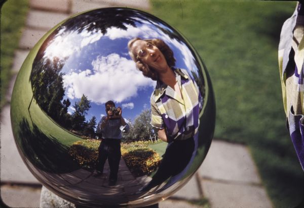 Reflection of Sid, holding a camera, and an unknown woman, standing in front of a gazing ball in a garden.