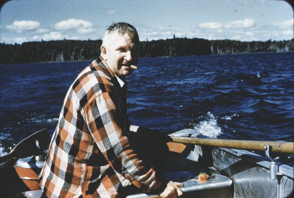 Sid sitting in a rowboat on a lake with a cigar in his mouth. Wearing an orange, white and black checkered shirt, he is turned sideways looking towards the camera. In the distance is the treed shoreline. 