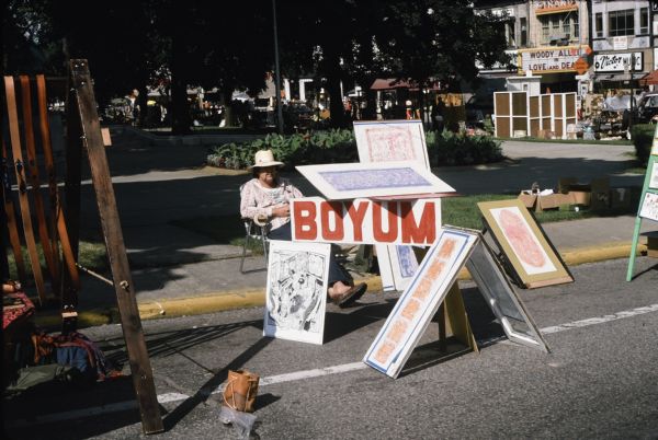 Display of Sid's art works on the street at what appears to be a fair on the Capitol Square. A man, possibly Steve Boyum, is sitting in a chair behind the display. Other vendors are in the background. The Strand Theatre on E. Mifflin Street is in the background. 