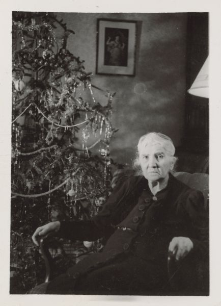 Sophie Smith (Mrs. Arthur) Pugh sitting in an arm chair next to a Christmas tree. She is wearing a jacket over a dark dress with large buttons. The tree is decorated with ornaments, and tinsel with beaded and chenille garlands.