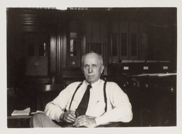 Arthur Pugh (1860-1948) posing while sitting in the office of the state treasurer in the Wisconsin State Capitol. He is holding his glasses and is wearing a white shirt, tie, and suspenders. Pugh, a native of Racine, worked as an accountant in the treasurer's office from 1900 until two months before his death in 1948.   