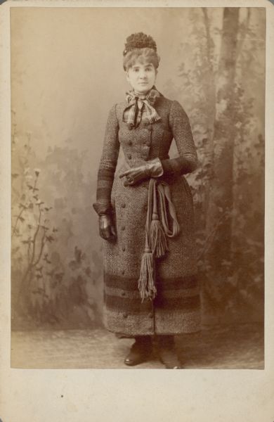 Full-length studio portrait of Sophie Smith (1861-1945), posing standing in front of a painted backdrop. She would later marry Arthur Pugh. She is wearing a full-length, double breasted winter coat with a tasseled sash, plaid scarf tied in a bow at the collar, earrings, leather gauntlet gloves, and a hat.