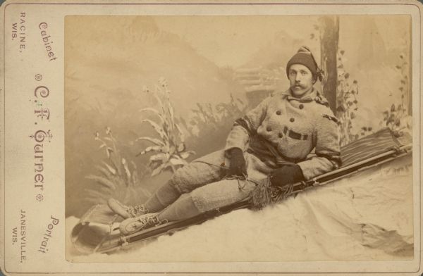 Portrait of a man wearing a stocking cap, mittens and a double-breasted coat reclines on a toboggan which is supported on a studio prop at an angle in front of a painted winter backdrop. He is also wearing knickers, long stockings, knitted gloves, and soft laced boots.