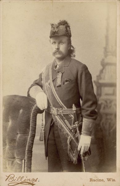 Three-quarter length studio portrait in front of a painted backdrop of an unidentified man standing and resting his arm on the back of an upholstered chair with a tassel. He is wearing the uniform of the Patriarchs Militant fraternal order, a branch of the Independent Order of Odd Fellows, which includes a plumed hat, sash, gauntlet gloves and chest ribbon. His left hand is resting on a lodge sword. 
