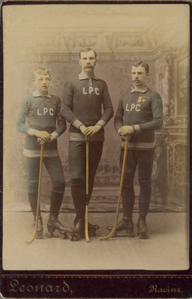 Full-length group portrait in front of a painted backdrop of three members of the Lakeside Polo Club for a cabinet card portrait, wearing roller skates and athletic uniforms. They are holding modified hockey sticks and have medals pinned to their shirts. They are, left to right: C. Smith, C. Stocking, and Arthur Pugh. Polo was an early name for roller hockey.