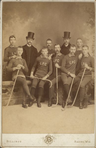 Group portrait in front of a painted backdrop of eight members of the Lakeside Polo [Roller Hockey] Club posing with two finely dressed gentlemen wearing coats and top hats. The club members are wearing athletic uniforms and five of them are holding modified hockey sticks.  Pictured are, left to right: J. Rowlands, E. Griswold, unidentified, C. Smith, P.S. Fuller, C. Stocking, unidentified, Arthur Pugh, Jackson I. Case, and W.A. Driver.  