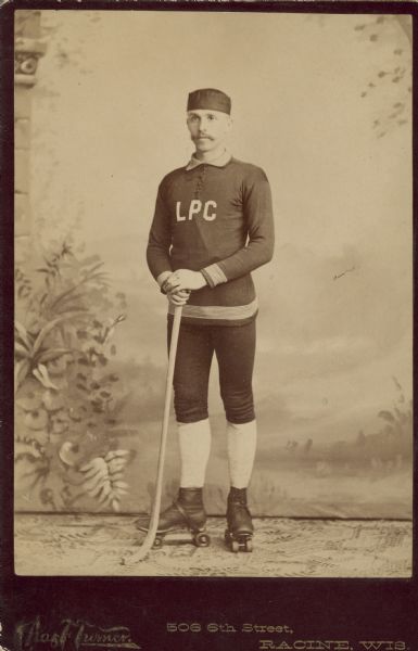 Full-length studio portrait in front of a painted backdrop of Arthur Pugh standing. He was the captain of the Lakeside Polo Club. He is wearing a cap, athletic uniform and roller skates, and is holding a modified hockey stick. "Polo" was an early name for roller hockey.