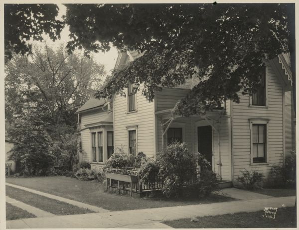 View from street towards the residence of Arthur and Sophia Pugh, 107 W. Gorham Street. Bridal wreath spirea bushes bloom around the open front porch, and there are bedding plants in a wooden planter beside the porch railing. The tidy one and one half story house has a bay window on the side. There are brackets under the porch roof and modest gable decoration. Two concrete tracks form a driveway on the left.