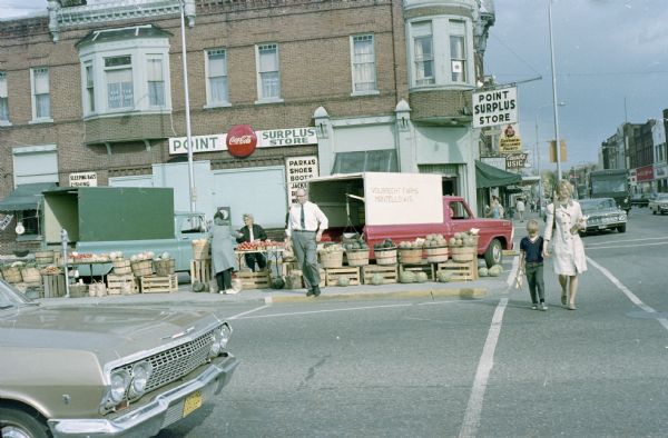 View from sidewalk of a food market across the street set up along a sidewalk. A woman and a child are walking across the street in the crosswalk. The Point Surplus Store is on the corner. The sign on the truck parked at the market reads: "Volbrecht Farms, Montello, Wis."