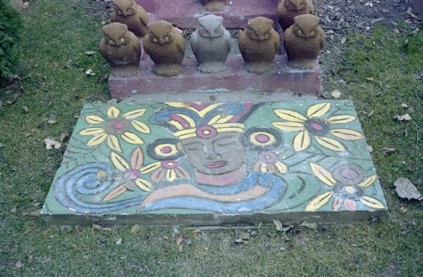 Painted low relief, set in the lawn near the base of the Buddha sculpture, of a Buddhist face, along with abstract flower patterns. Owls are set along the base of the Buddha sculpture, which is not shown.