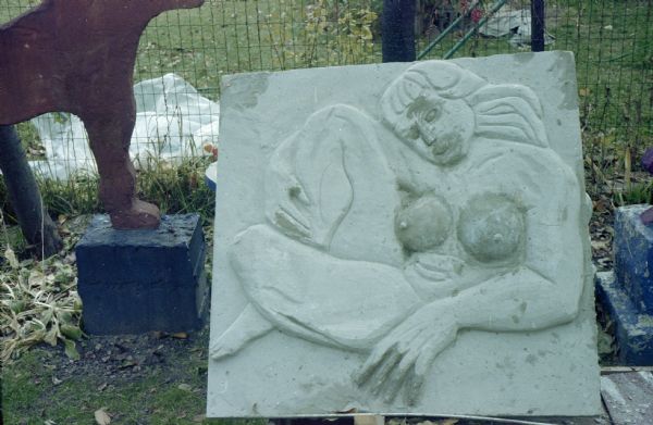 Unfinished large rectangular concrete-slab bas-relief of a nude woman identified as "Diana." She has long hair and bangs, and sits in a relaxed pose. Supported by her left elbow, her left leg wraps around the right at the ankle, with her right hand falling below the knee. The sculpture measures 45" x 41" x 14". The white plastered slab is leaning against a wire fence in Sid's backyard.