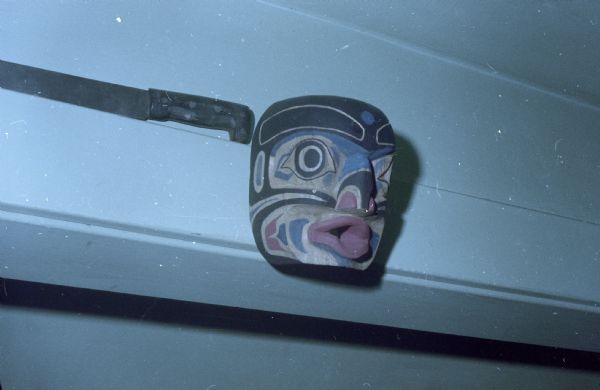 Wooden carved mask inspired by Pacific West coast native art styles with an eagle beak and eyes, gaping fish mouth, and fish tail motifs on its cheeks. It is painted in blue, red, white and black. It is hanging in Sid Boyum's house over a doorway. A knife is resting on the door frame.  