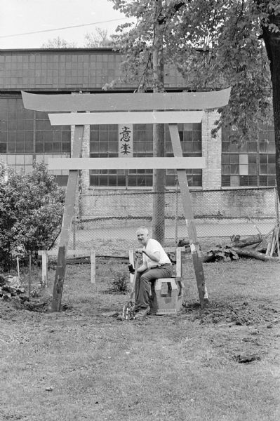 View towards backyard with Sid is sitting on a stool under the torii gate that has recently been anchored into the ground. He has a pitchfork. Madison-Kipp Corporation is in the background behind a chain-link fence.