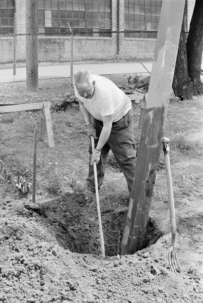 Sid is digging a hole in the ground for the torii gate he is installing in his backyard. Madison-Kipp Corporation is in the background behind a chain-link fence.