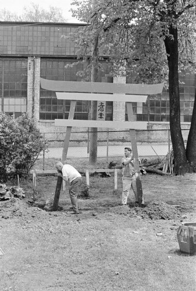 Sid and a man erecting the torii gate in Sid's backyard. Madison-Kipp Corporation is in the background behind a chain-link fence.