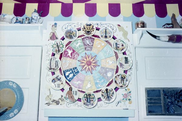 A decorative clock with roman numerals on plates, signs of the zodiac, and other illustrations displayed on the kitchen wall of Sid's house. 