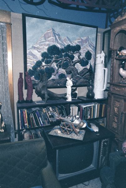 View of a corner of Sid's living room. There is a painting by Sid of a pine tree in a mountainscape hanging on the wall. Below it is a bookshelf with several sculptures and other ceramic art objects on it. On top of the television is a cannon in the shaped of an imaginary dragon. On the bottom right, Sid can be seen lying on the couch with a cigar in his mouth.