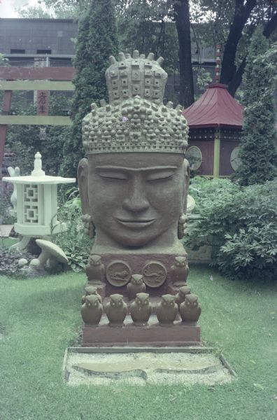 Large Cambodian-styled, cast concrete "Monumental Oriental Head" of a Buddha painted red in Sid's backyard with heavily textured light-colored hair and topknot. With dimensions of 110 x 45 x 78 inches, it sits on a thick base ornamented with medallions bearing symbols of the Zodiac. Figural owls sit beneath the bust on the base and in the space in front. 
