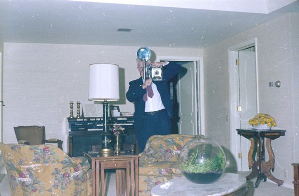 Sid looking through a 4 x 5 Busch Pressman camera toward the viewer; a flashbulb is mounted to the side. He stands in a living room furnished with upholstered bucket chairs, nested wooden end tables, tall lamp, and marble top coffee table. There is a terrarium on a table in the foreground, and an organ in the background with two brass candlesticks.