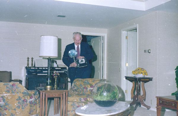 Sid checking his 4 x 5 Busch Pressman camera toward the viewer; a flashbulb is mounted to the side. He stands in a living room furnished with upholstered bucket chairs, nested wooden end tables, tall lamp, and marble top coffee table. There is a terrarium on a table in the foreground, and an organ in the background with two brass candlesticks.  