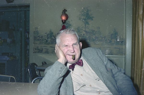 Portrait of Sid wearing a suit. He is making a funny face and has his right eye closed while smoking a cigar. 
