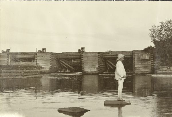 Ernest Copeland standing on a rock while fishing near Gordon Dam. He is wearing pajamas and a hat.
