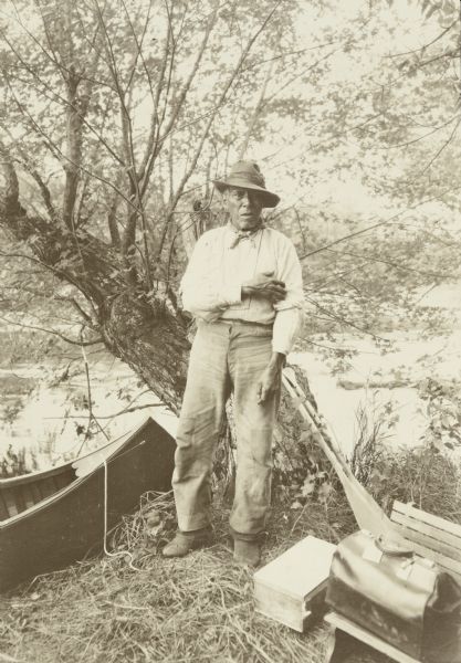 Bill Williams standing in front of a tree at the shoreline. There is a canoe next to him on the left, and paddles, bags and boxes are on the ground on the right.