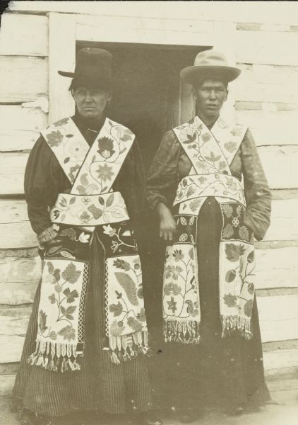 Two people, probably Ojibway, posing together wearing hats and elaborately beaded garments, with blouses with puffy sleeves. They are standing in front of the doorway of a building with wood siding.