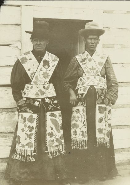 Two people, probably Ojibway, posing together wearing hats and elaborately beaded garments, with blouses with puffy sleeves. They are standing in front of the doorway of a building with wood siding. 