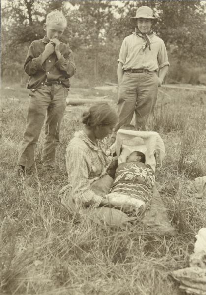 A young woman is sitting on the ground holding her infant in a cradleboard? Carl (left) and Clay are standing behind her.