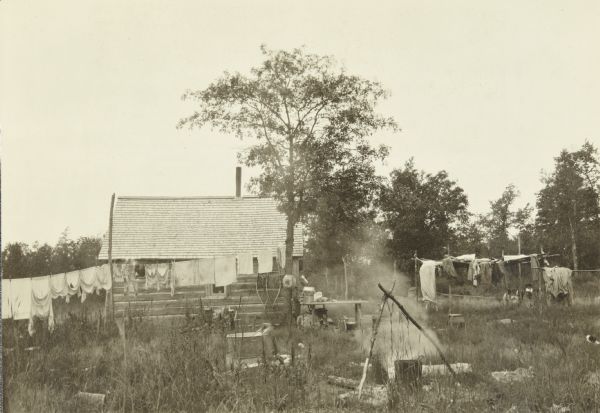 View of a house and yard. There is laundry hanging out to dry and, in the foreground, a cooking pot over a fire.