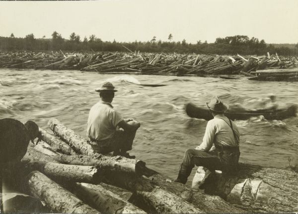 Two men watching from the shore as Bill Marr shoots the Kettle Rapids in a canoe. The spectators are sitting on logs.